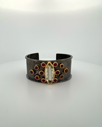 Luxury cuff bangle crafted in black rhodium plated 925 sterling silver with thick 18K gold vermeil. Featuring a marquise green amethyst centerpiece surrounded by garnet gemstones. Adjustable statement bracelet for 16–17 cm wrists.