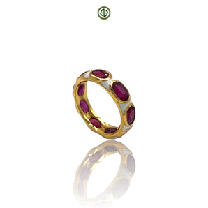 A gold-colored eternity ring with red ruby stones set in a white enamel band, displayed on a reflective surface.