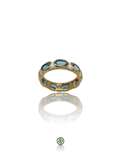 A gold-colored eternity ring with blue topaz stones set in a smooth white enamel band, displayed against a white background with a reflective surface.