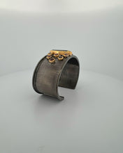 Luxury cuff bangle crafted in black rhodium plated 925 sterling silver with thick 18K gold vermeil. Featuring a marquise green amethyst centerpiece surrounded by garnet gemstones. Adjustable statement bracelet for 16–17 cm wrists.
