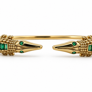 Gold-coloured open bangle with two facing alligator heads, textured scales, green baguette-cut stones and round green eye details