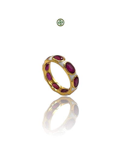 A gold-colored eternity ring with red ruby stones set in a white enamel band, displayed on a reflective surface.