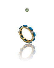 A gold-colored eternity ring with white enamel and turquoise inlays, displayed on a reflective surface.